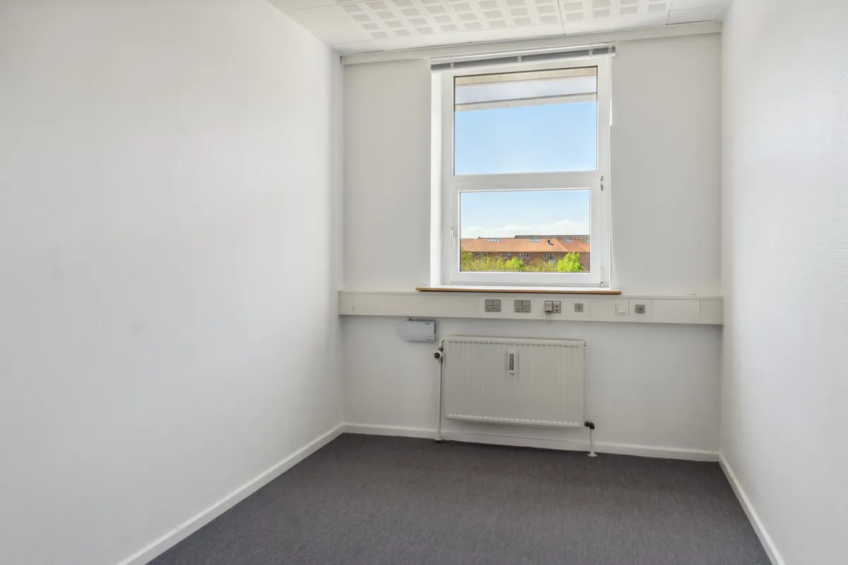 En tom kontorværelse med hvide vægge, hvor vinduet er åbent og viser bygninger og grønne træer i baggrunden