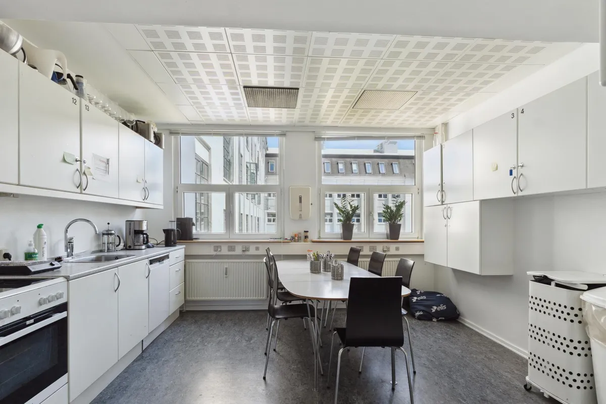 Kontor med lyse, hvide vægge og møbler samt vinduer med udsigt til et bygningskompleks og planter.