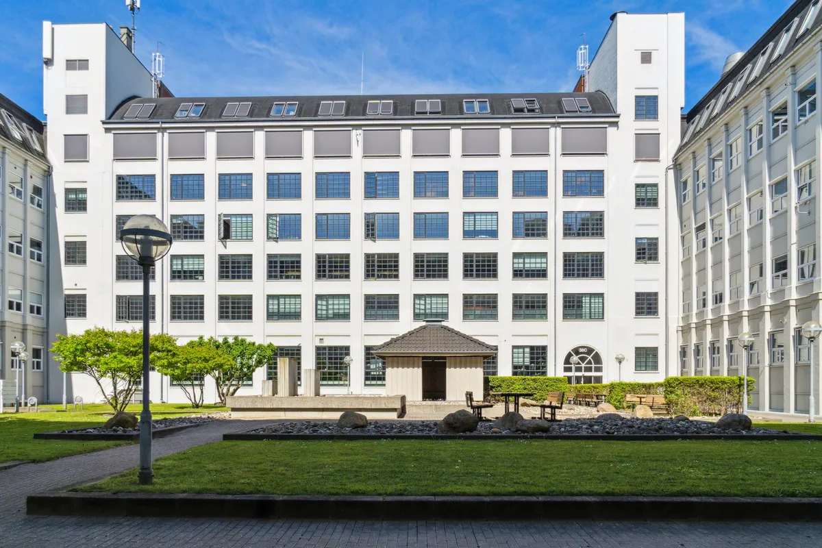 Moderne kontorbygning med flere gulve og vinduer, solpaneler på taget og en lille have med træer og græs.