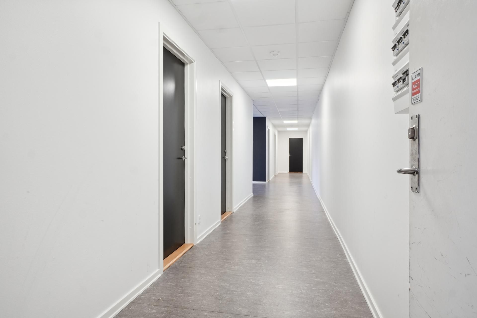 Kort gang mellem hvide vægge med højloft og lyse plader med døre og skilte