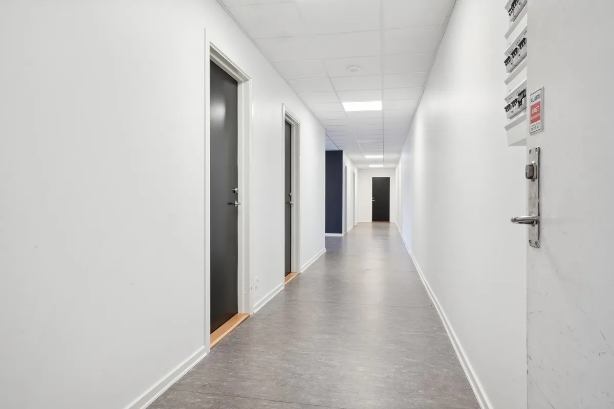 Kort gang mellem hvide vægge med højloft og lyse plader med døre og skilte