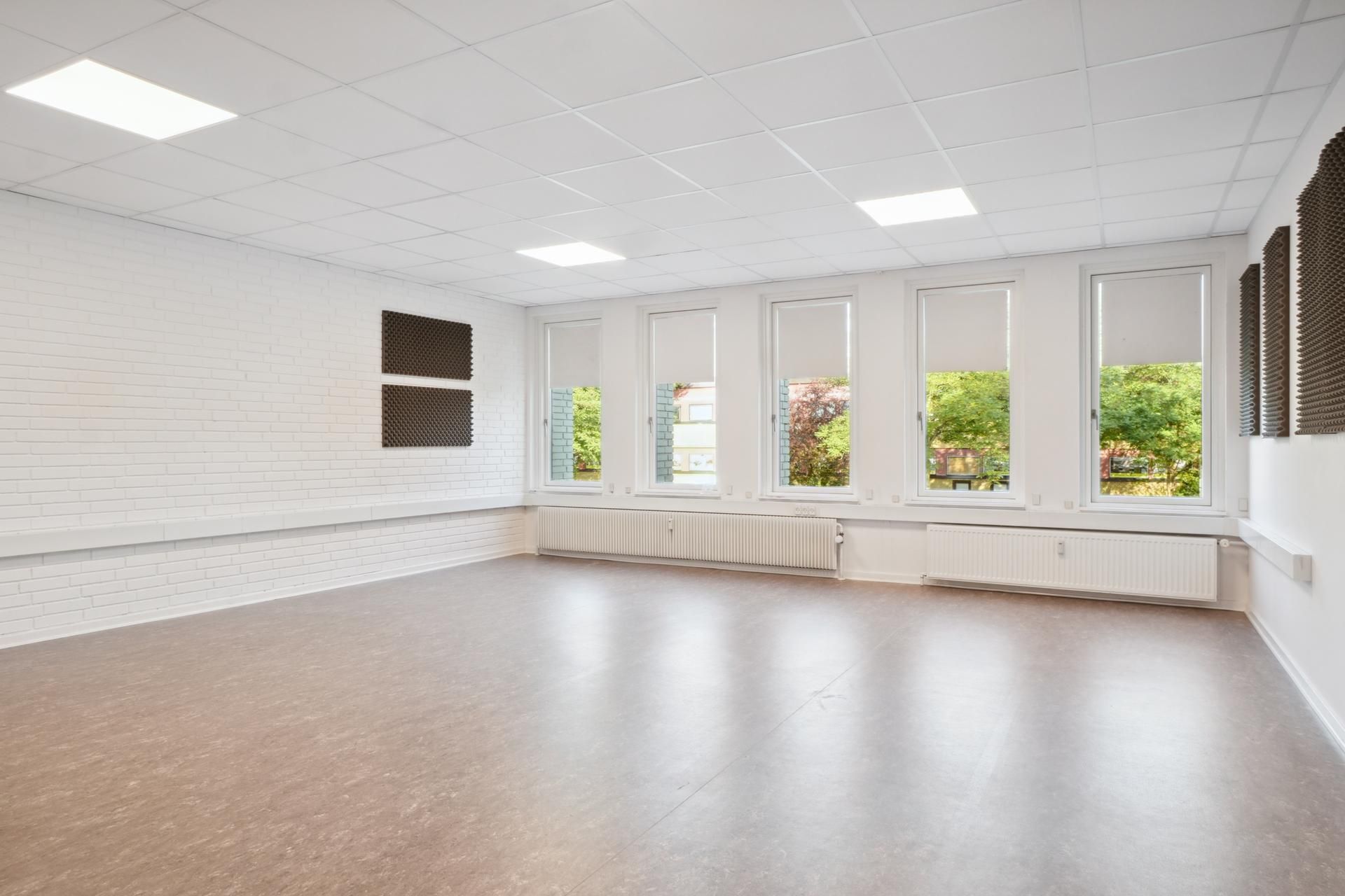 En stor, lys og rummelig lokale med hvide vægge, lysegule lofter, trægulve og vinduer med gardiner og radiatorer.