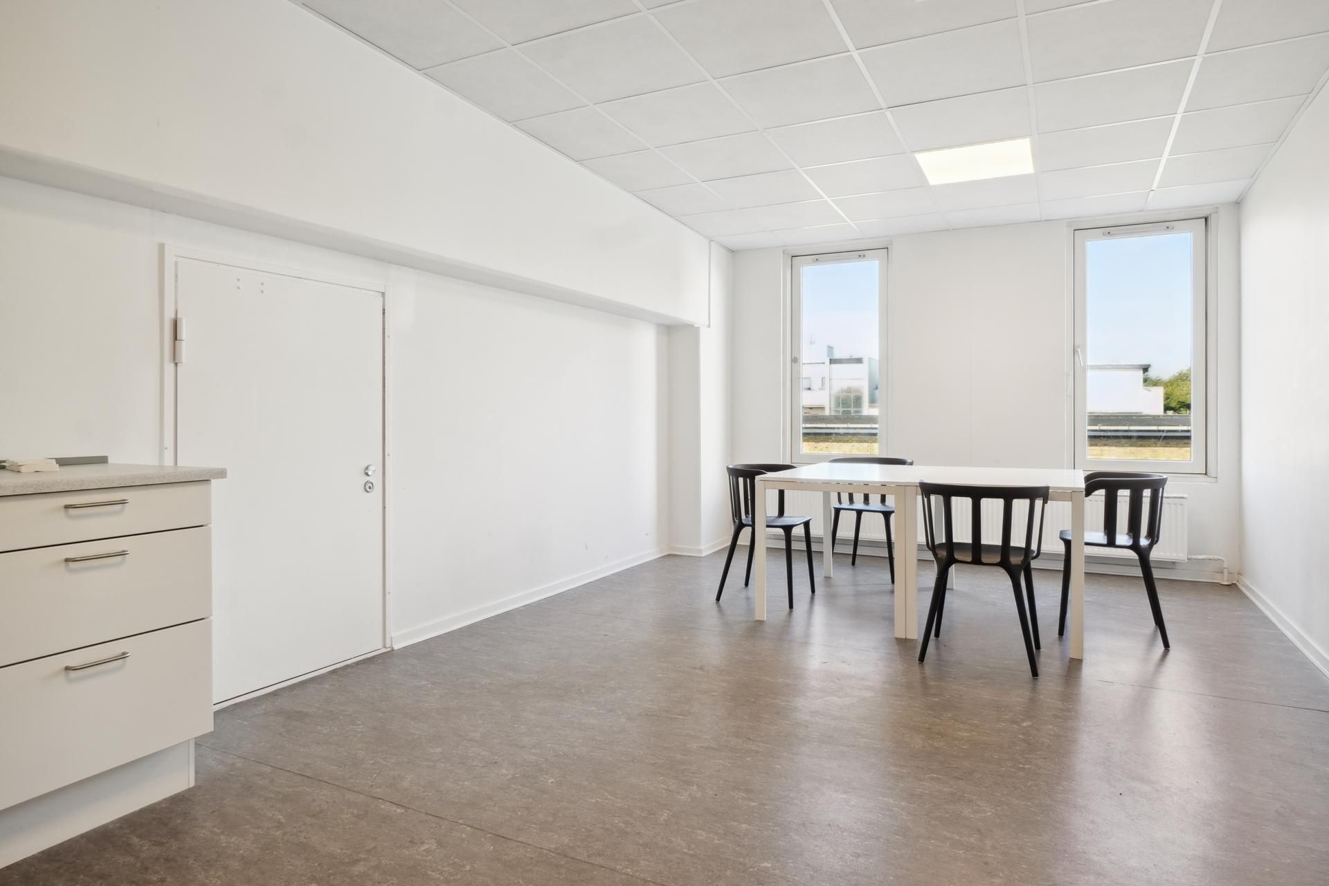 Moderne rum med hvide vægge og vinduer, trægulv og et lille hvidt bord med fire stole samt en hvid kommode med tre skuffer