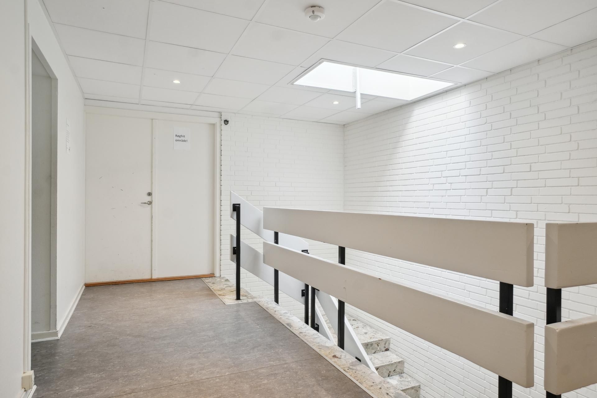 Øvelseslokale med hvid pudsede vægge, højloft, trapper, stålgesims og indgangsdør med skilt med tekst "Regnfri omride!"