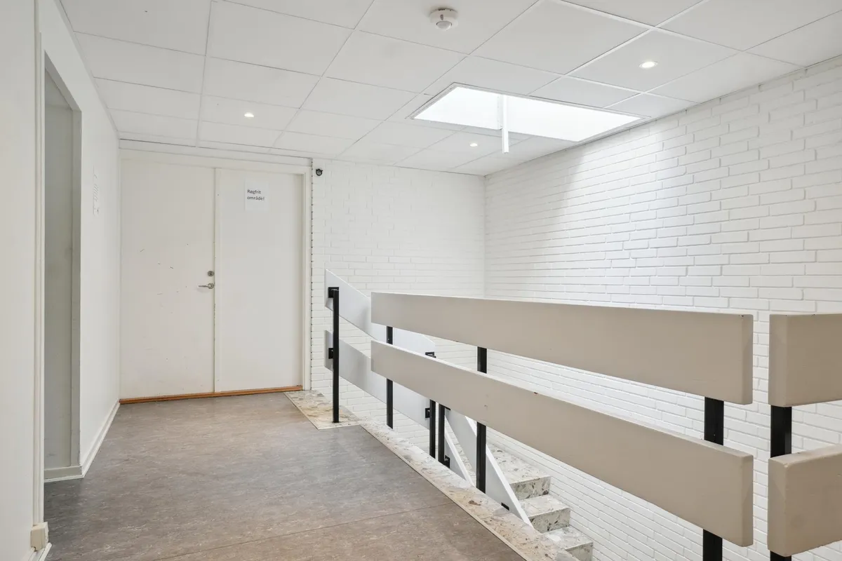 Øvelseslokale med hvid pudsede vægge, højloft, trapper, stålgesims og indgangsdør med skilt med tekst "Regnfri omride!"