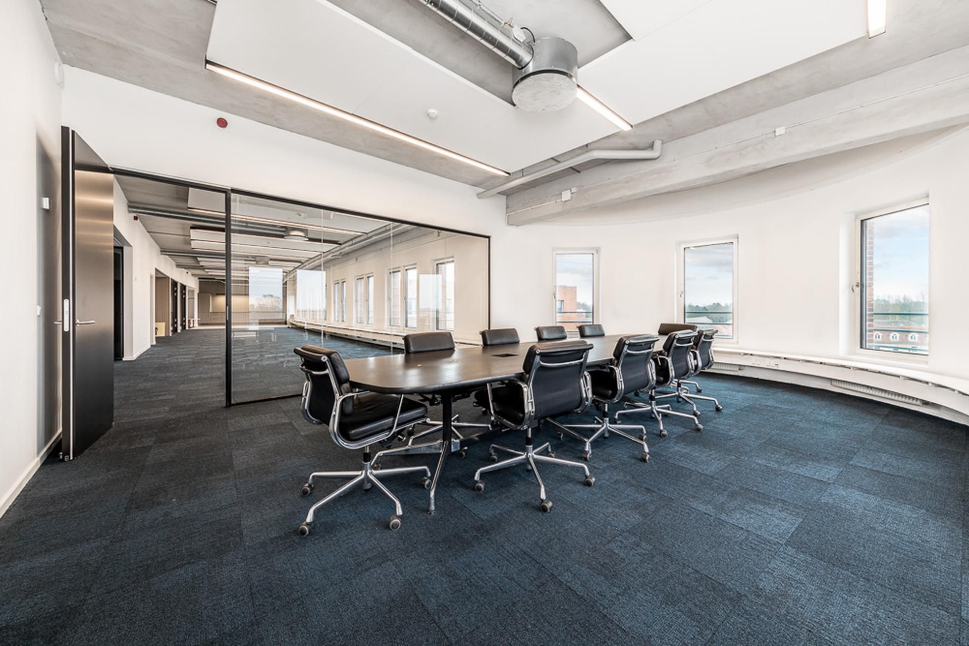 Et stort mødelokale med en lang bord og flere stole rundt om bordet samt vinduer langs væggen