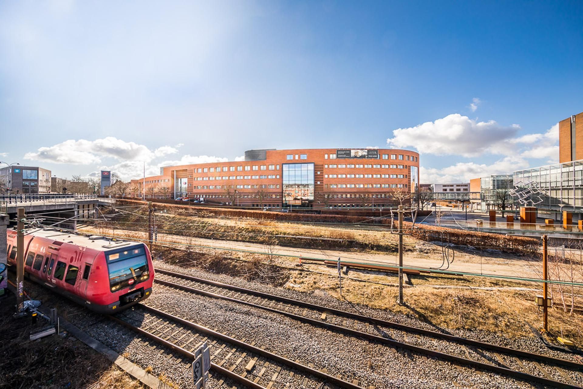 Et moderne bygningskompleks med flere etager og glasvinduer. En togskinne med en rød togvogn i solskin. En bro i baggrunden