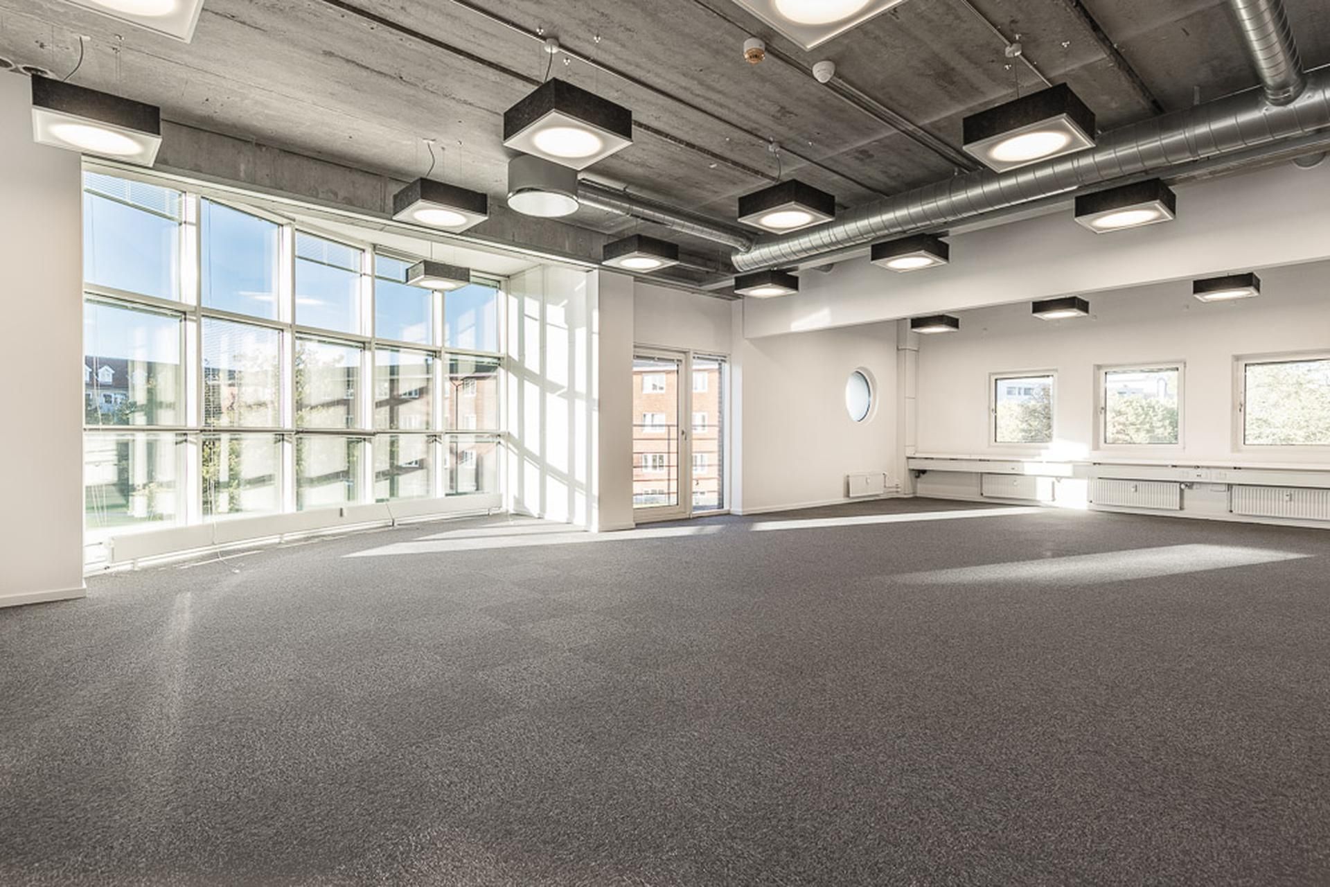 Et stort, moderne kontor med store vinduer, højt loft og lyse loftslampe samt grågulvt gulv.