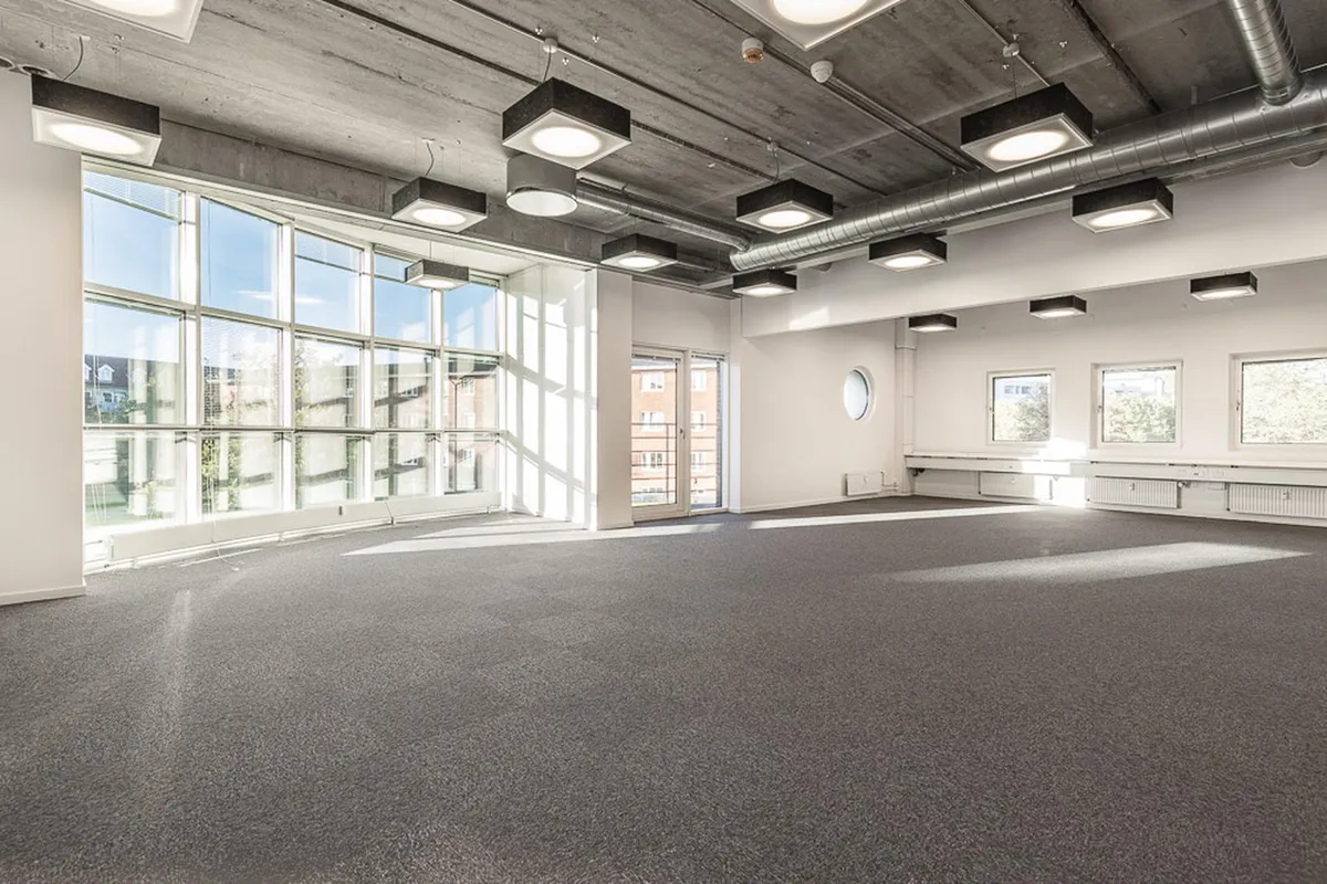 Et stort, moderne kontor med store vinduer, højt loft og lyse loftslampe samt grågulvt gulv.
