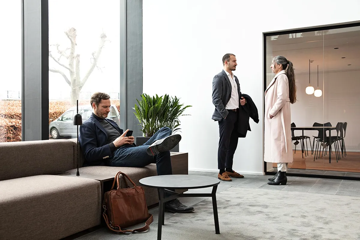 To mænd og en kvinde samles og snakker i et moderne kontorlokal med lyse vinduer, hvor man ser en plante og en sofa i