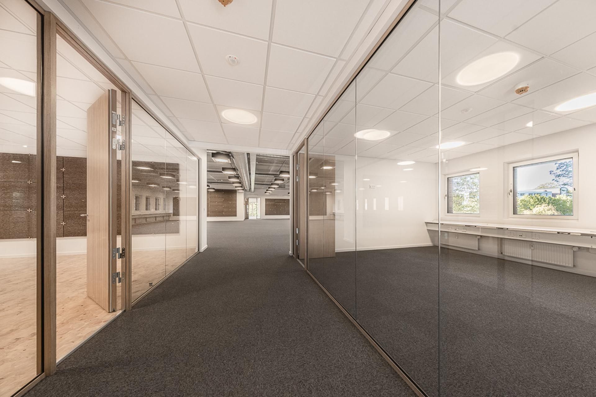 Modernt kontorlokal med store glasvægge og hvide lofter. Væggene er lyse og reflekterer godt. Gulvet er dækket med gråt