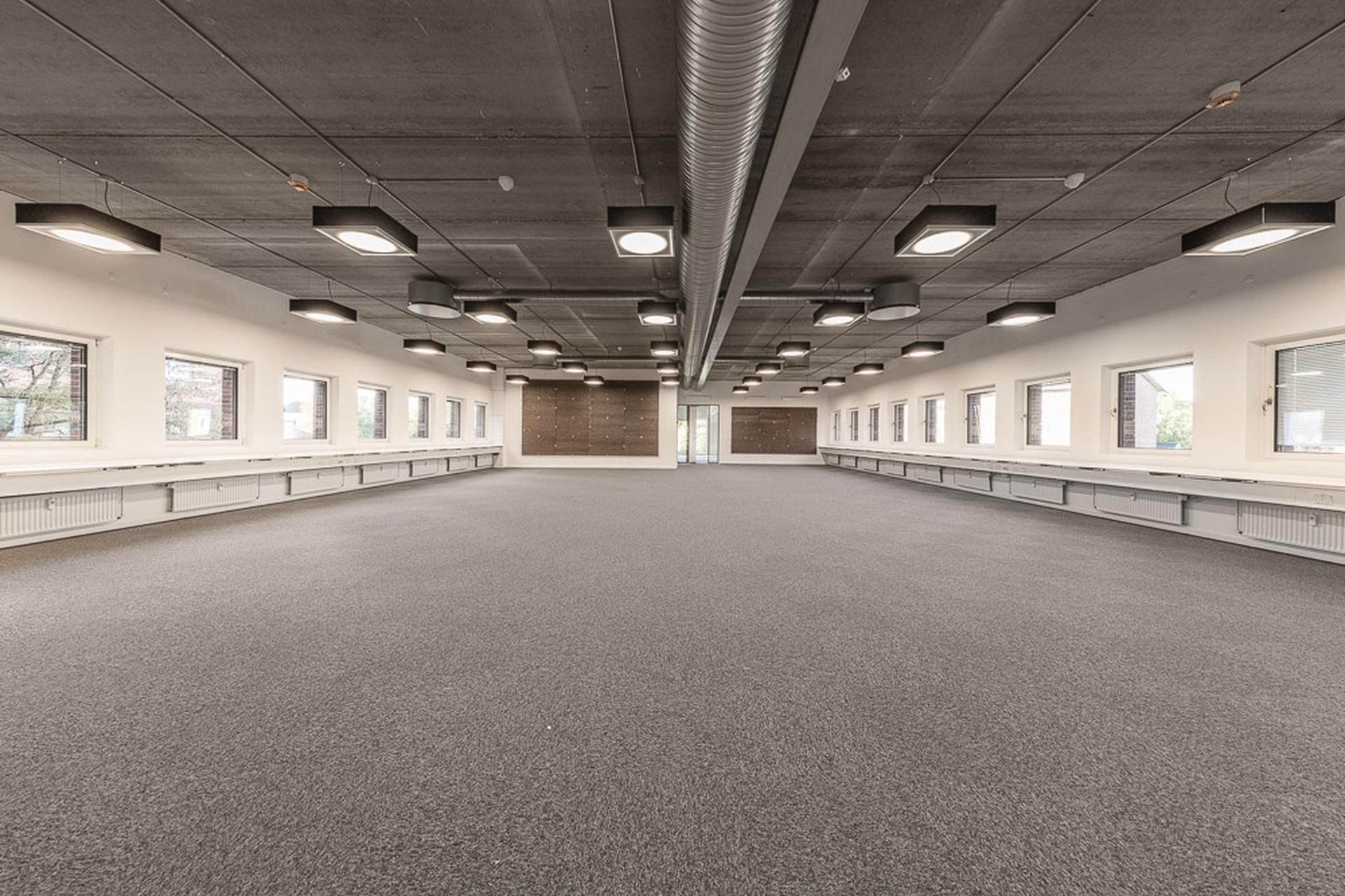 Kontor med lyse loftslampe og store vinduer i Kgs. Lyngby, idet man kan se et tomt gulv og møbler langs væggen