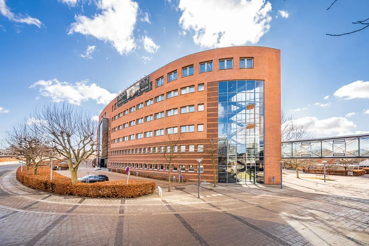 Kontorbygning med glas og røde mursten i Kgs. Lyngby med skur og gitterbro. Solskin og skyer i baggrunden og skov med træer