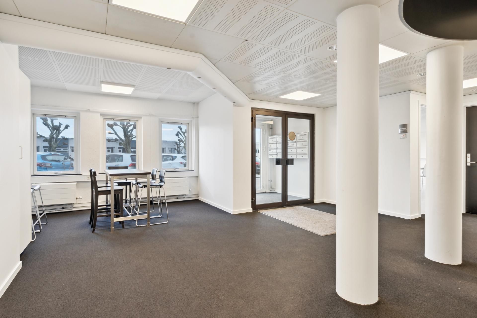 En tom kontorrum med hvide vægge, gulv og loft, træbord og stole samt fire vinduer med udsigt til parkeringsplads og biler