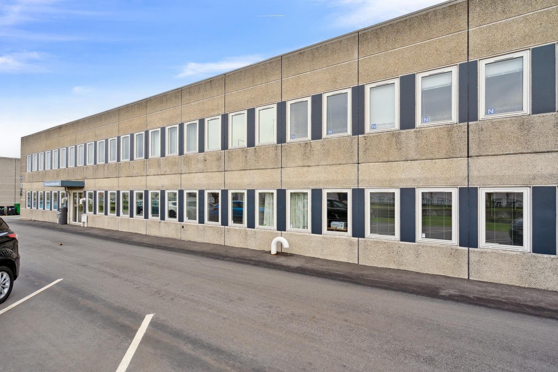 Betonbygning med vinduer og skygger i baggrunden, hvor der er parkeret biler og skraldespande