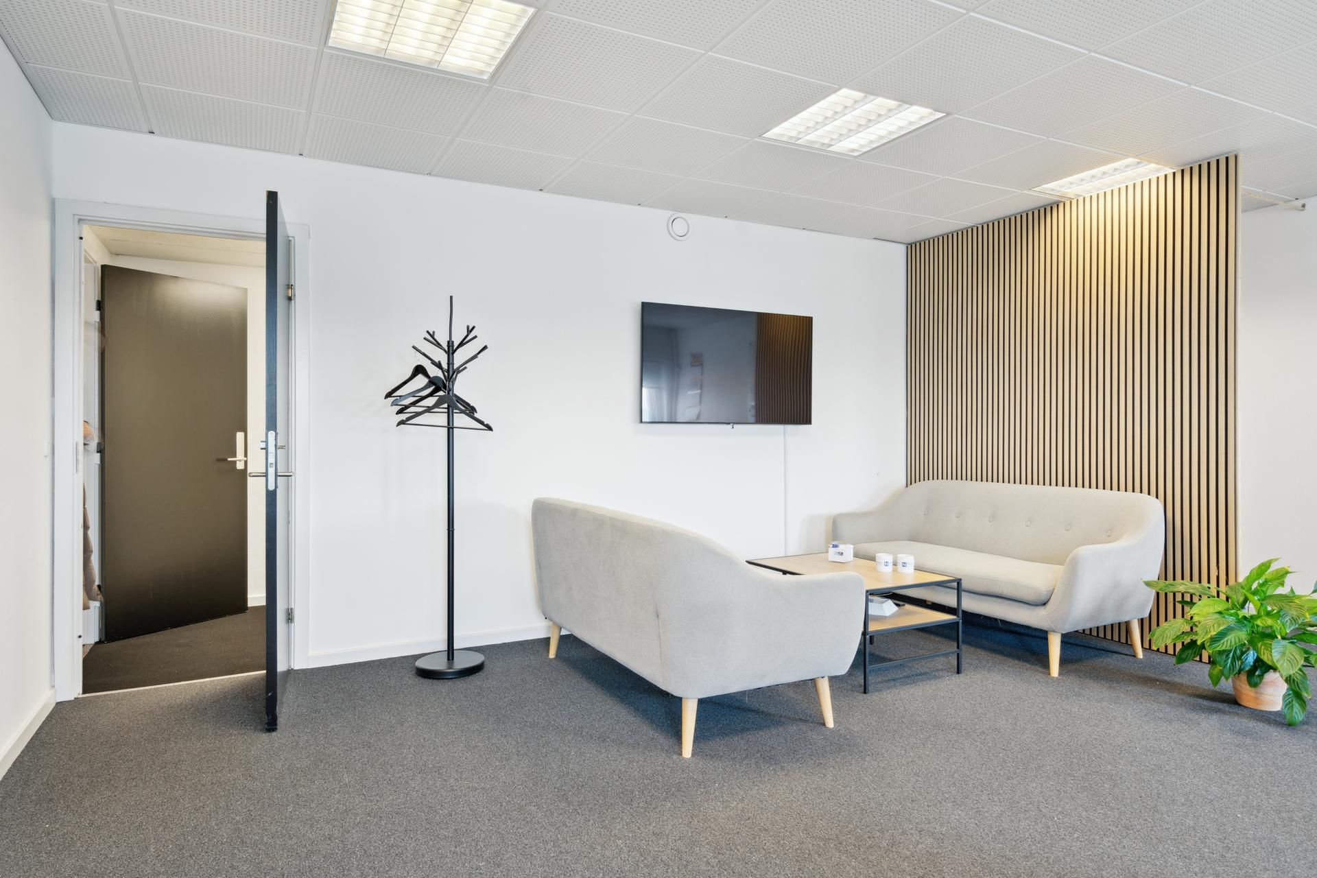 Et lille og rent kontor med hvide vægge og møbler, trægulv og plante i hjørnet, sammen med to lyse grå sofaer og et bord med