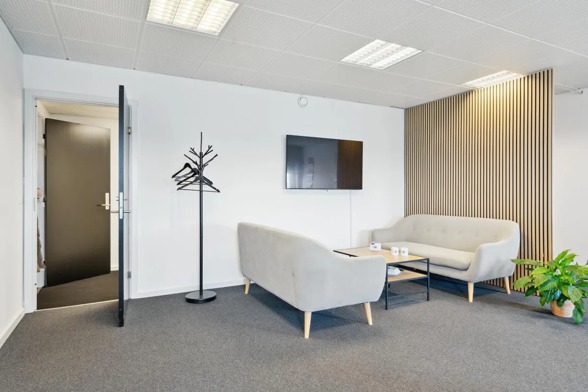 Et lille og rent kontor med hvide vægge og møbler, trægulv og plante i hjørnet, sammen med to lyse grå sofaer og et bord med