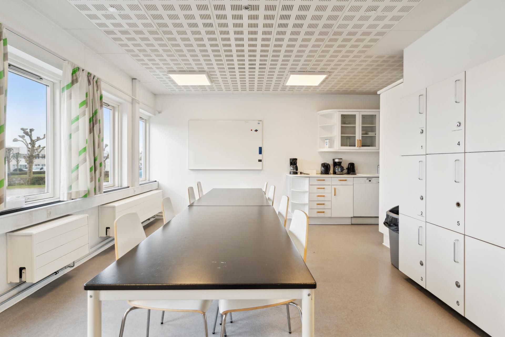 En minimalistisk stue med en lang sort bord, hvid trægulv og hvide vægge, hvor der er et hvidt whiteboard og lysegrå