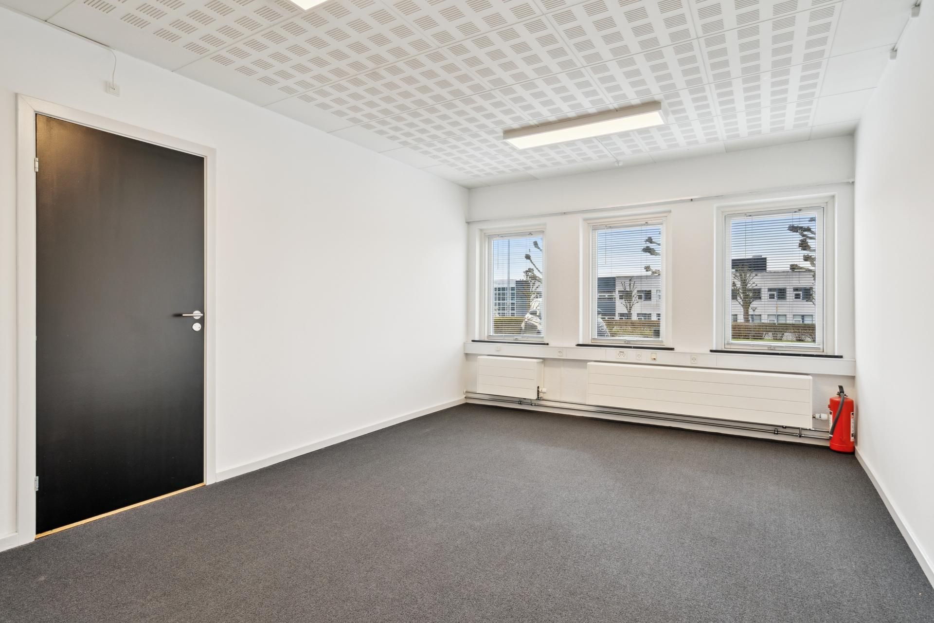 Tomt kontorrum med hvid væg og loft, gulv med grå tekstilbelægning, tre vinduer med gardiner og radiatorer samt en dør med