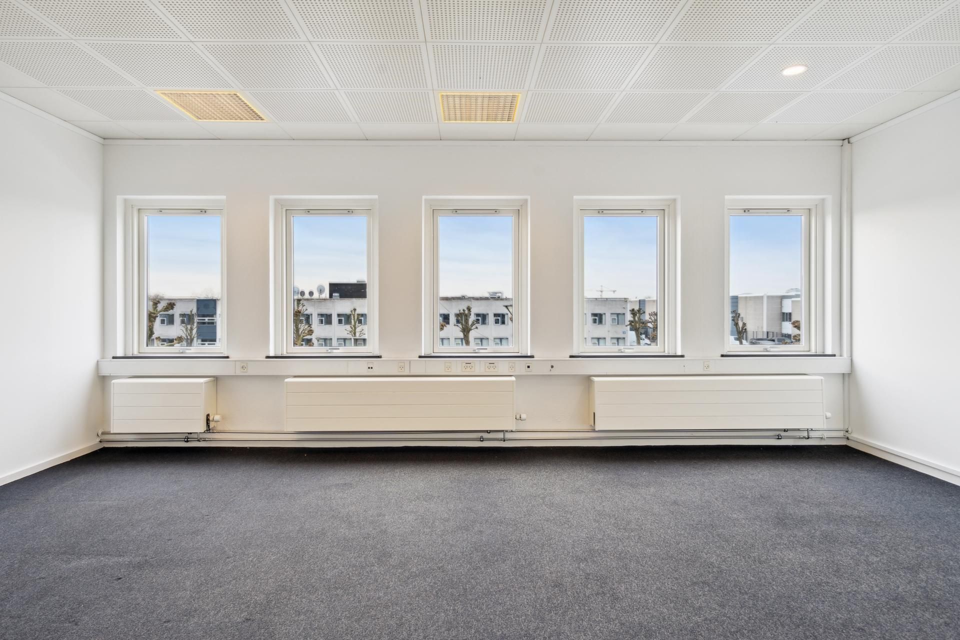 En tom kontorbygning med hvide vægge og loft, fire store vinduer med udsigt til bygninger og træer, og tre hvide radiatorer