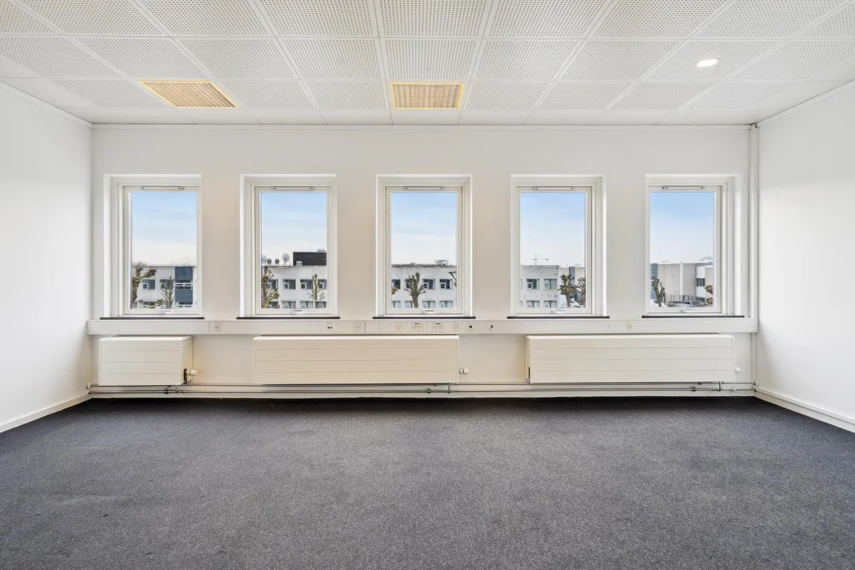 En tom kontorbygning med hvide vægge og loft, fire store vinduer med udsigt til bygninger og træer, og tre hvide radiatorer