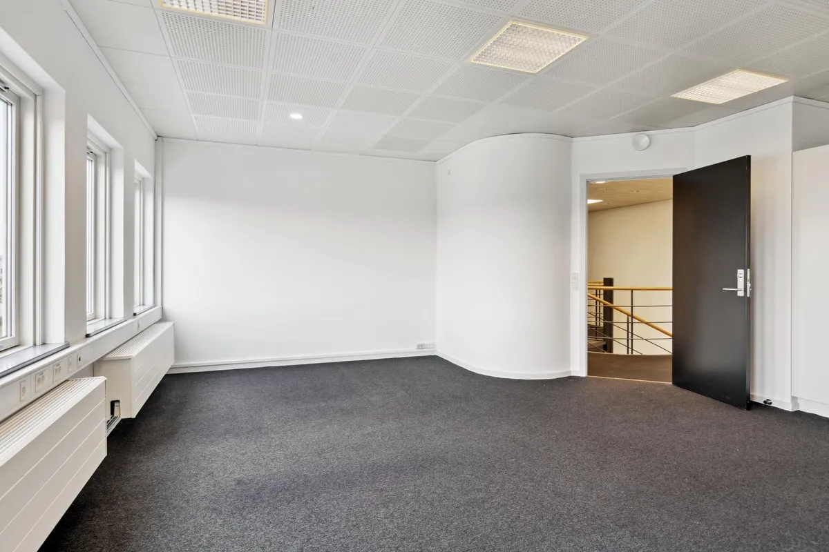Et tomt kontor med hvide vægge, grå gulvbelægning, store vinduer, åben dør, højloft, radiatorer og et trappeforløb