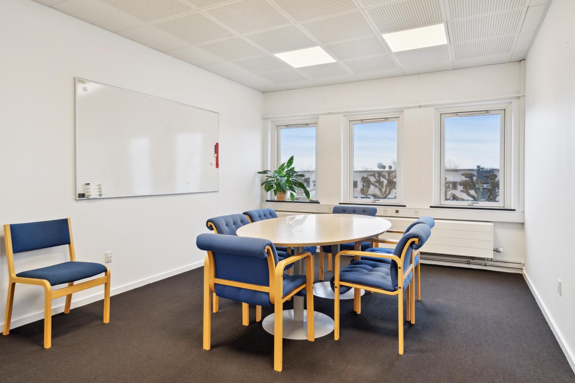 En mødelokale med lyse vægge og gulvtæppe, hvor der er en rund bord med flere blå stole omkring. Der er også en hvid