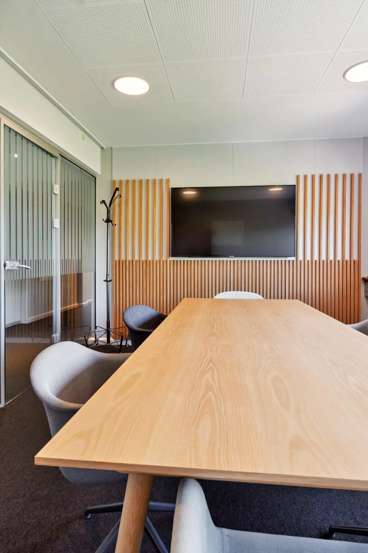 Kontorrum med lyse vægge og møbler samt en stor træbord og en stor tv-skærm på væggen