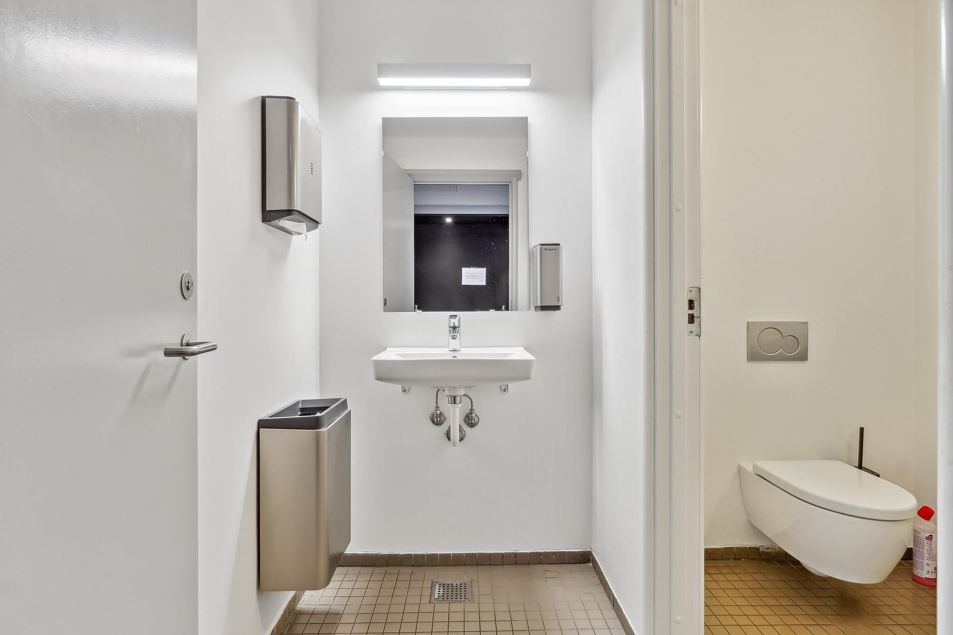 Hvidt badeværelse med håndklædeholder, håndvask, toalett, og samme farve på væggene og gulvet. To håndklædeuddeler er