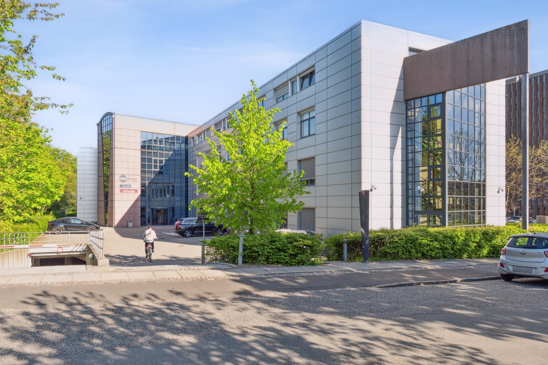 En person cykler forbi et kontorbyggeri med mange vinduer, hvor der er parkeret biler udenfor