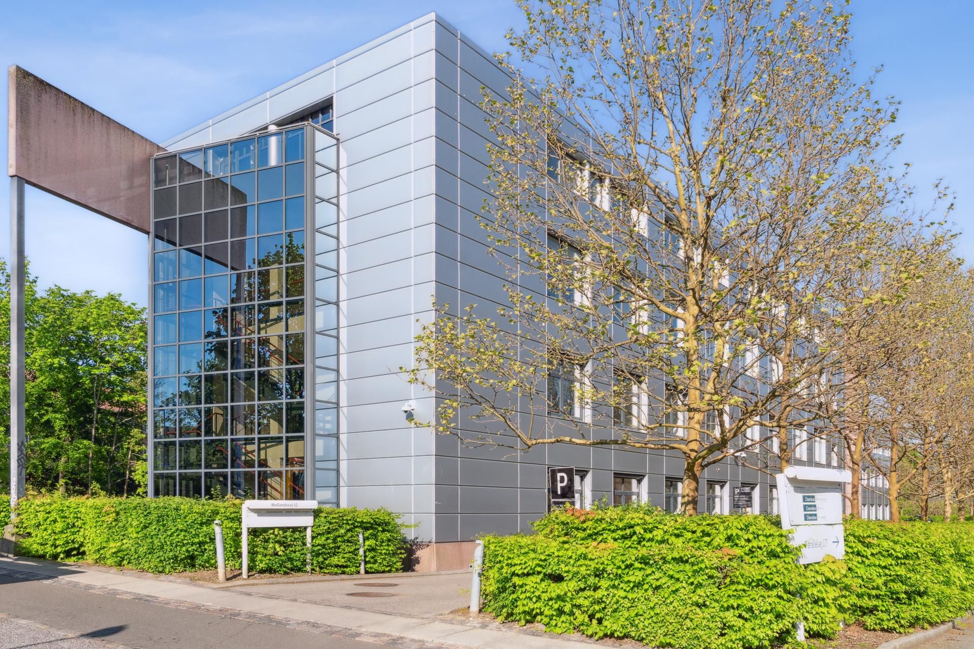Kontorbygning med glasvinduer og buske. Der er en skilt med bogstavet P og tallet 12. Der er en skygge fra et træ.