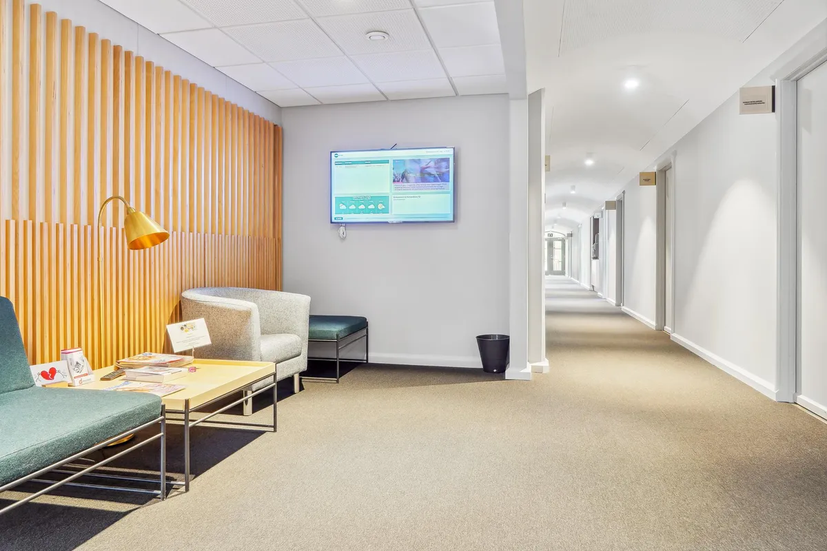 Kontor med lyse møbler, gul stanglampe, sofa, lænestol, gulvtæppe, og tekst på en monitor med hjertebillede på stolbordet