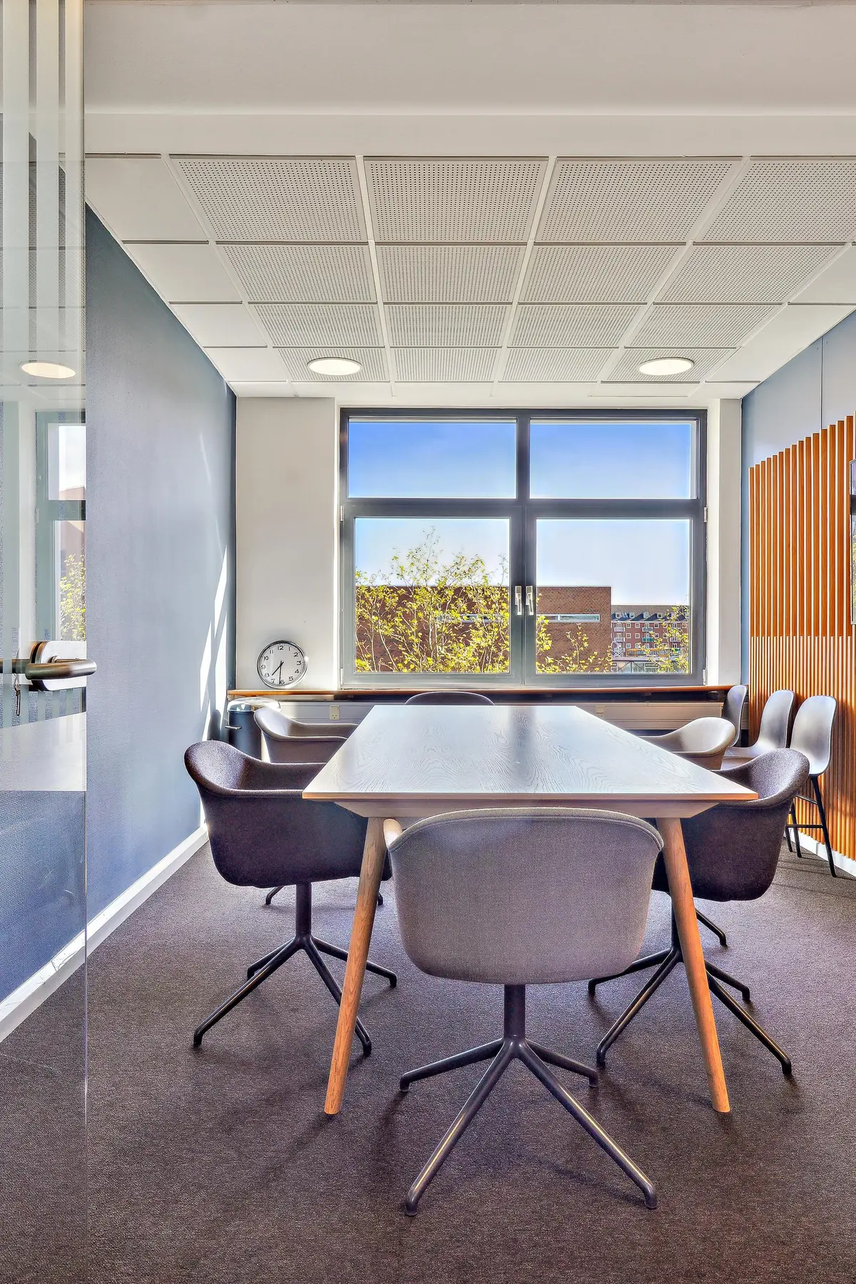 Kontor med lyse vægge, træpaneler, møbler og vinduer med udsigt til grønne træer og bygninger i baggrunden