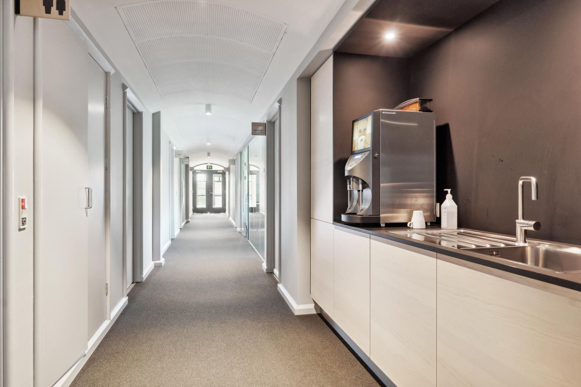 En lang kontorhvile med hvide vægge, gulv og loft. Køkken med flere hvide skabe, en kogeplade og en kaffemaskine. Der er en