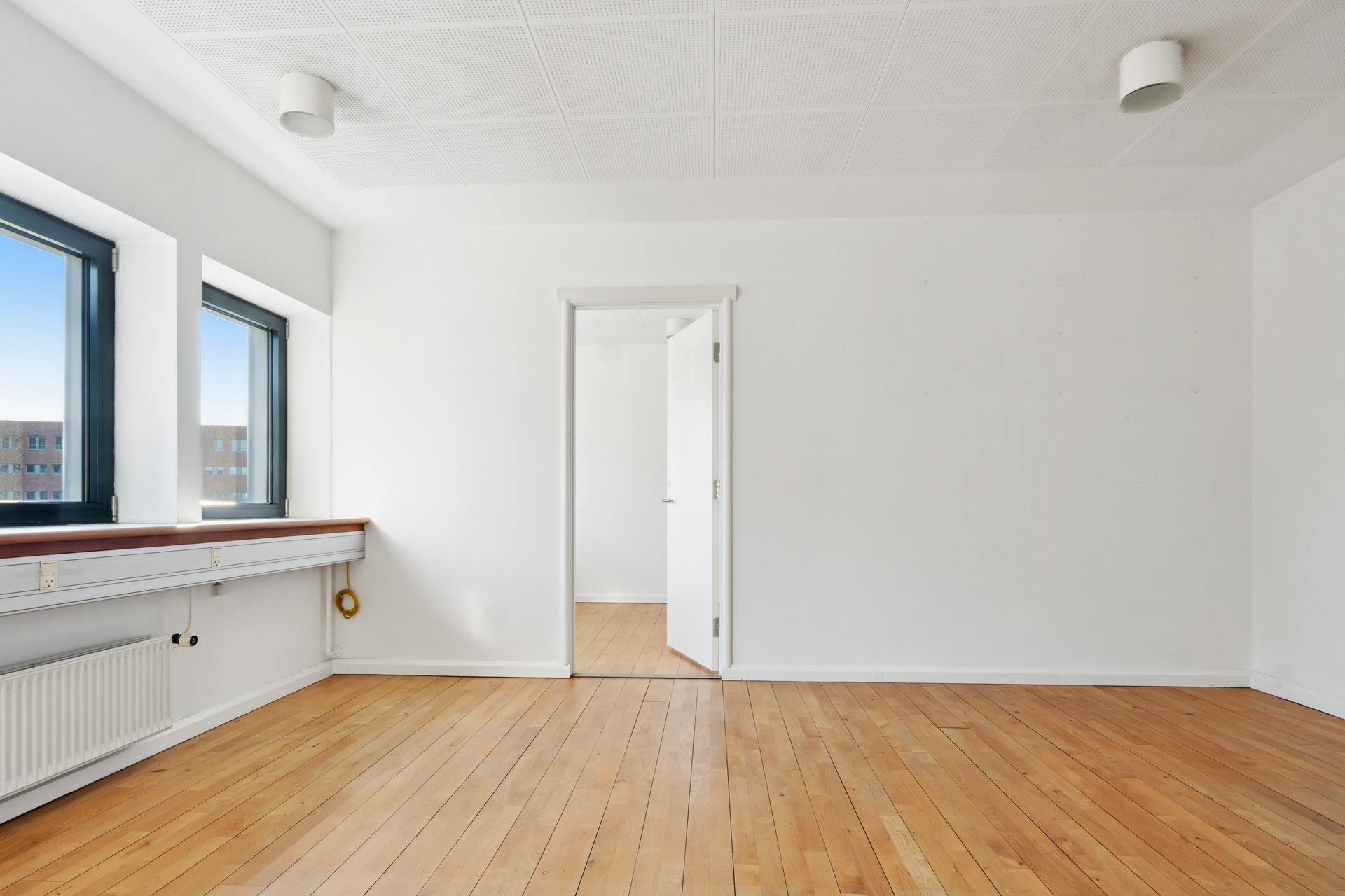 En stor, tom stue med høje lofter, hvide vægge og lyse trægulve samt to vinduer med udsigt til bygninger i baggrunden.