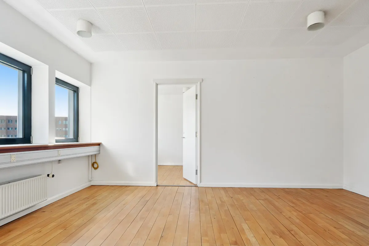 En stor, tom stue med høje lofter, hvide vægge og lyse trægulve samt to vinduer med udsigt til bygninger i baggrunden.