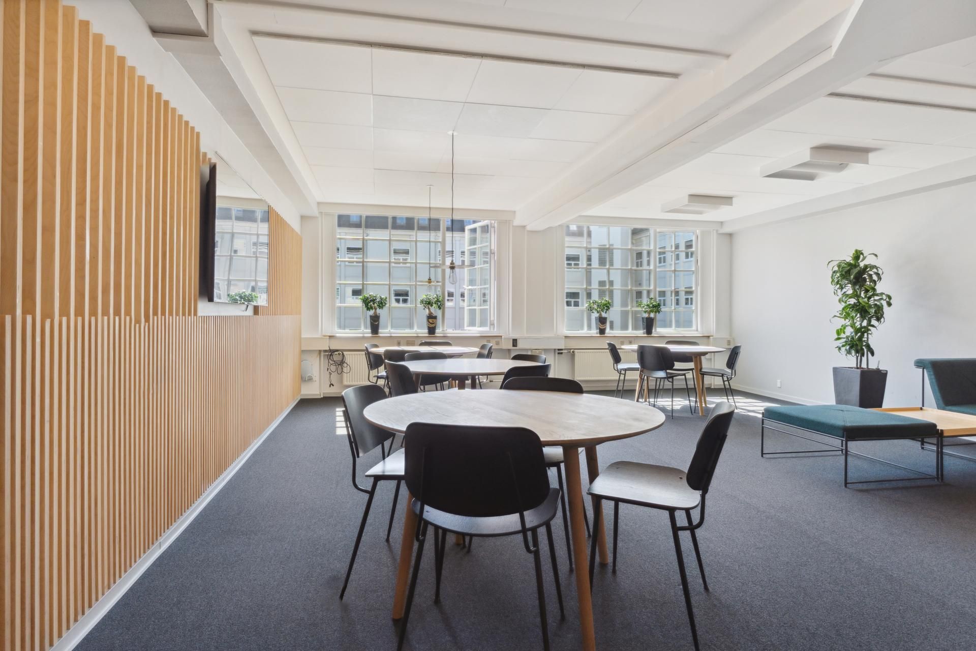 Kontorområde med møbelgrupper, plantepoter, og store vinduer på Østerbro. Moderne design med møbler og træpaneler.