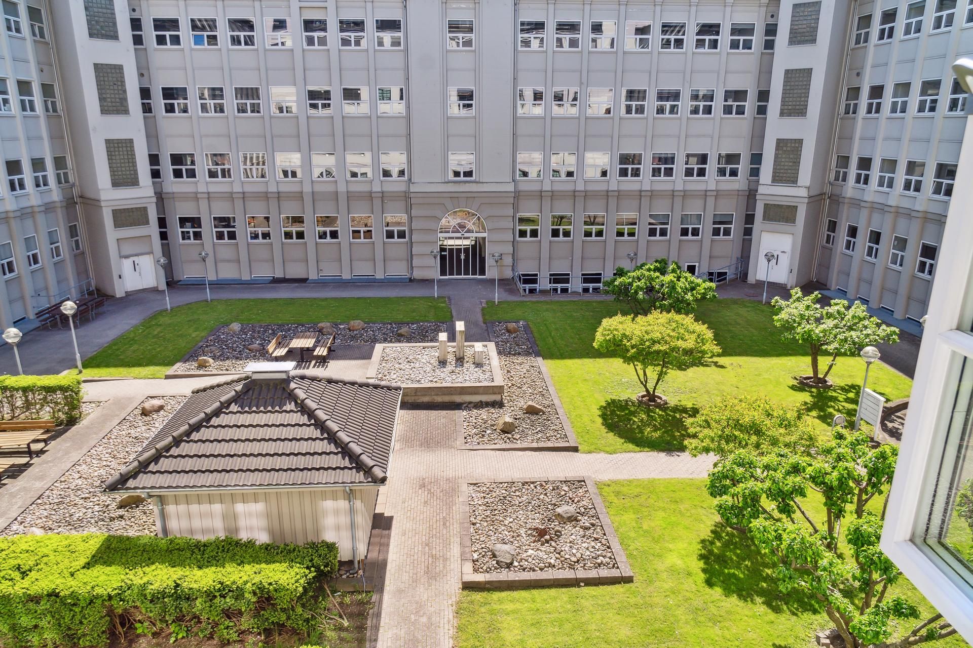 En gårdplads med grønne græsplæner og buske samt et pavillon i baggrunden og store kontorbygninger med glasvinduer