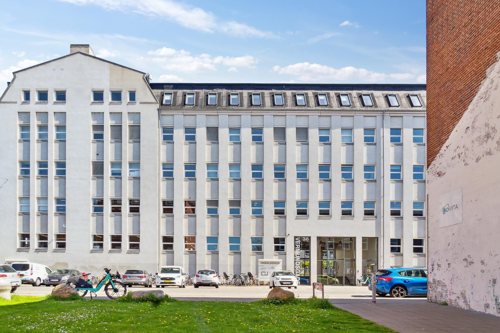 Kontorbygning med glasruder og skrånende tag med vinduer. Biler og cykler parkeret foran. Et skilt med teksten BoVita er