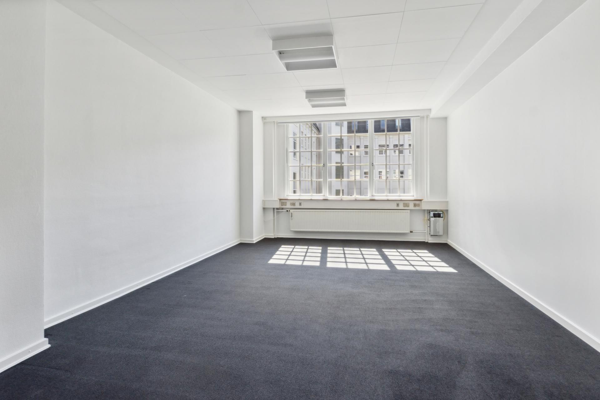 Kontorrummet er hvidt, med lysegrå gulvtæppe og glasruder med gardiner. Væggene er hvide, og der er to lysekroner og et