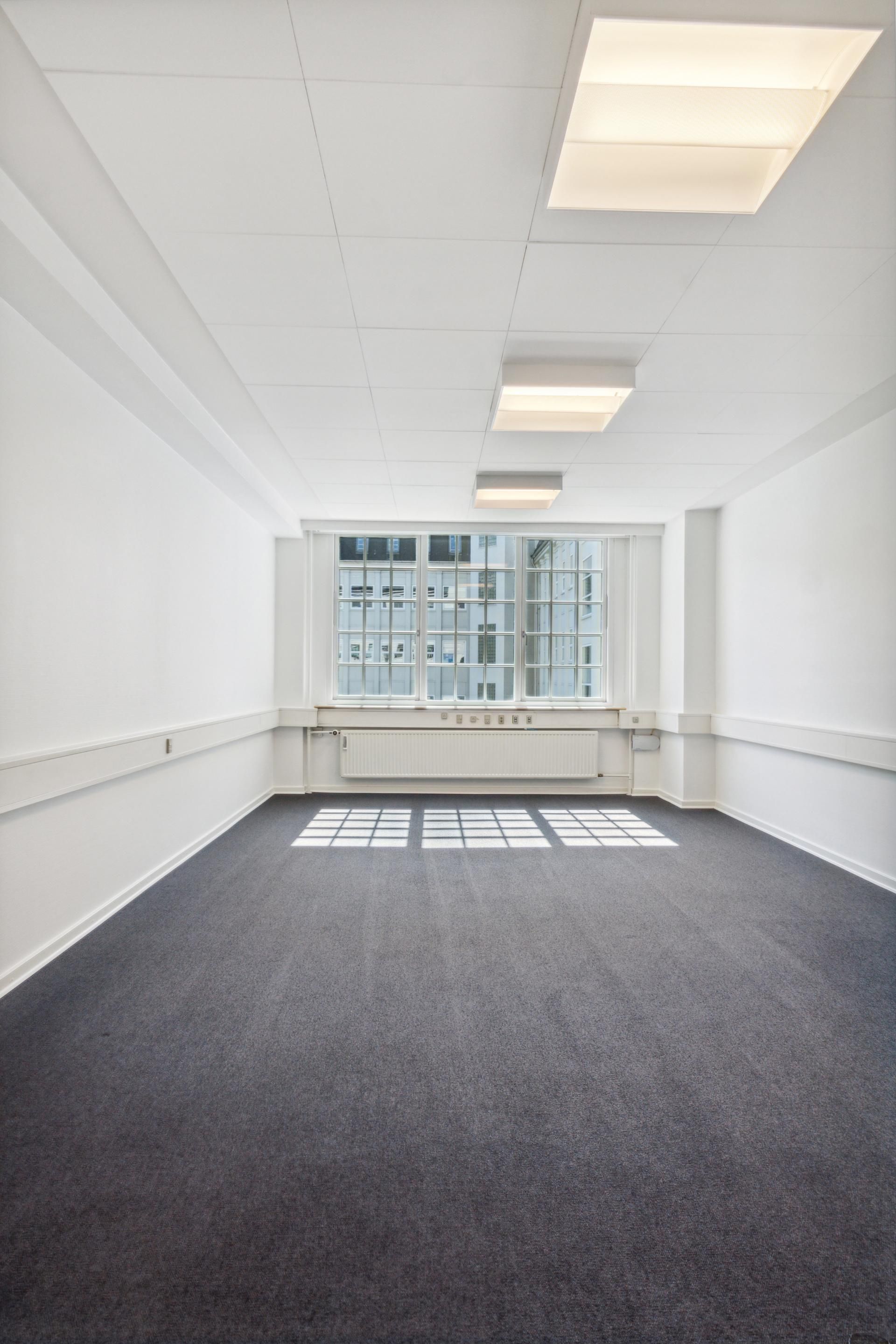 En tom og ren kontorlokal med hvide vægge og gulv, store vinduer og en hvid radiator. Loftet er hvidt med fire indbyggede