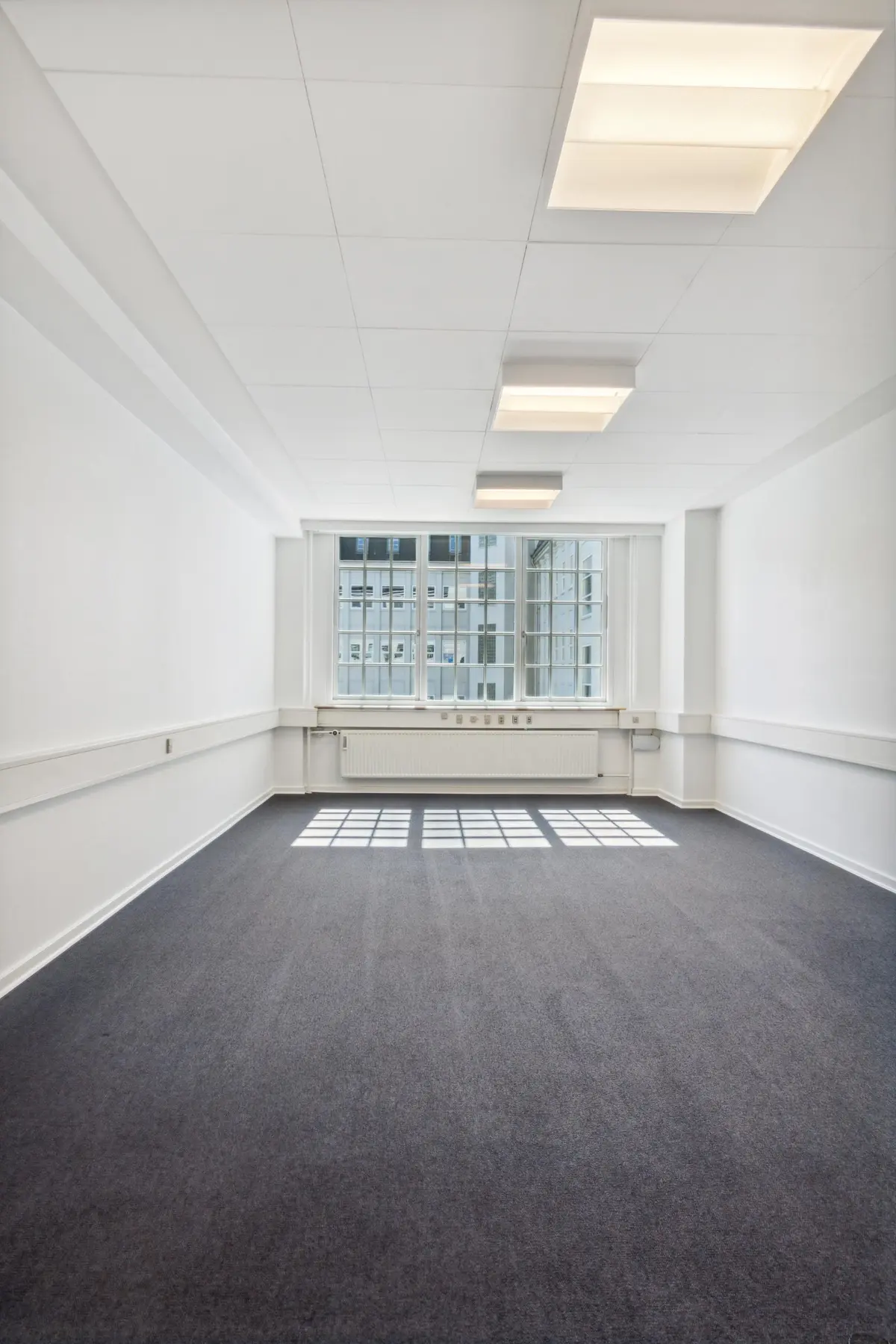 En tom og ren kontorlokal med hvide vægge og gulv, store vinduer og en hvid radiator. Loftet er hvidt med fire indbyggede