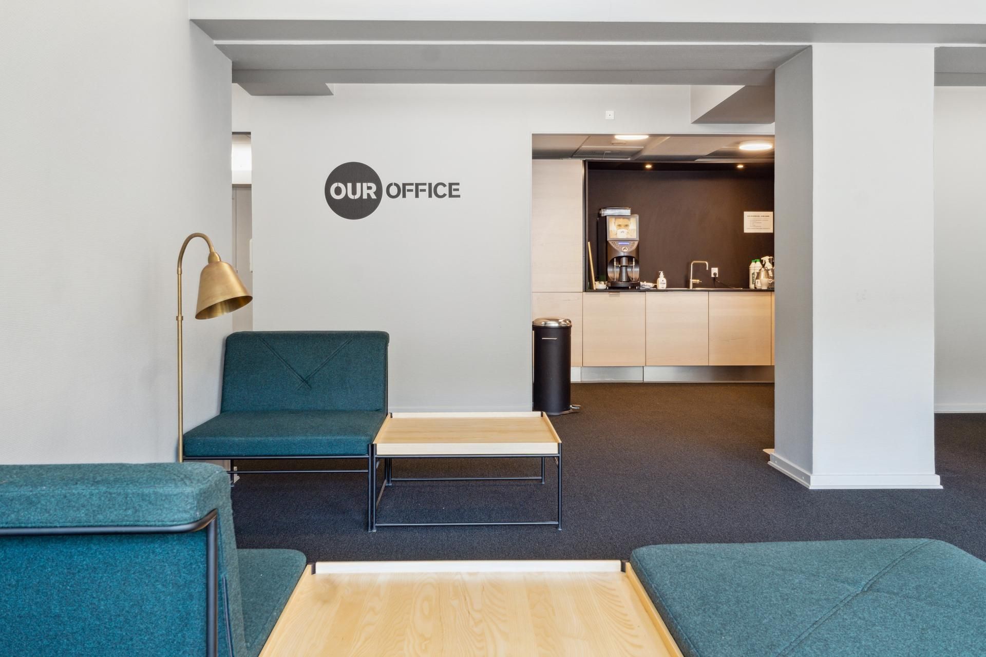 Kontorfællesskab med grønne lædersæder og lyse vægge, et kaffemaskine og en høj søjle i baggrunden