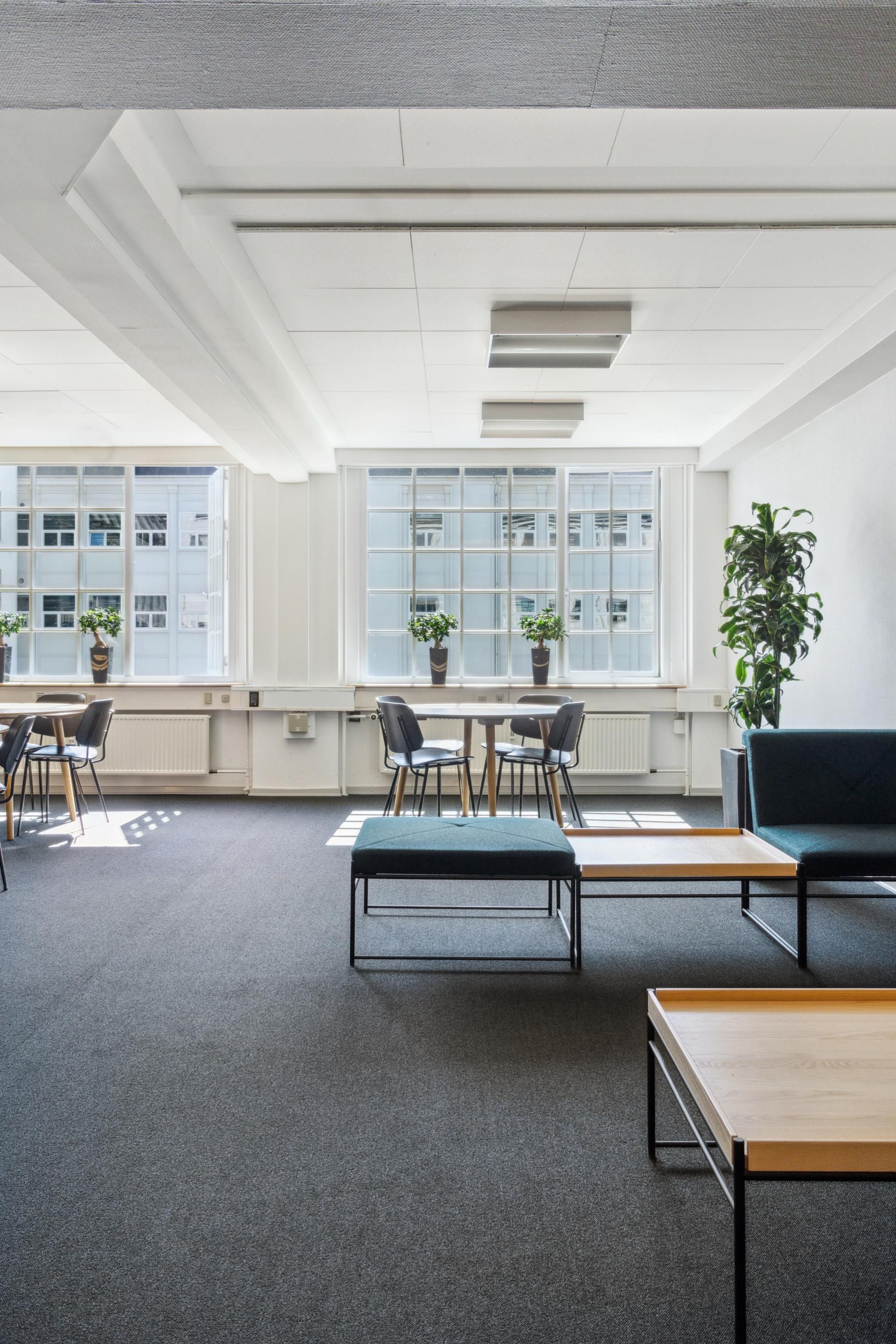 Moderne kontorlokaler med lyse vinduer, møbler og plante i hjørnet, samt to bordopsætninger med stole og en sofa.