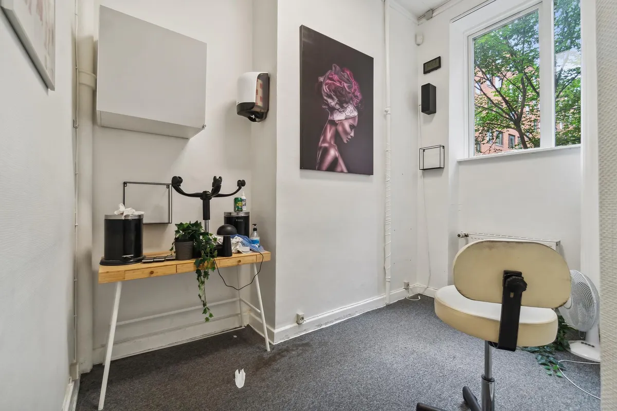 I et lille rum med hvide vægge og gulvtæppe er der et bord med en plante og et sæde med en ventilator til siden