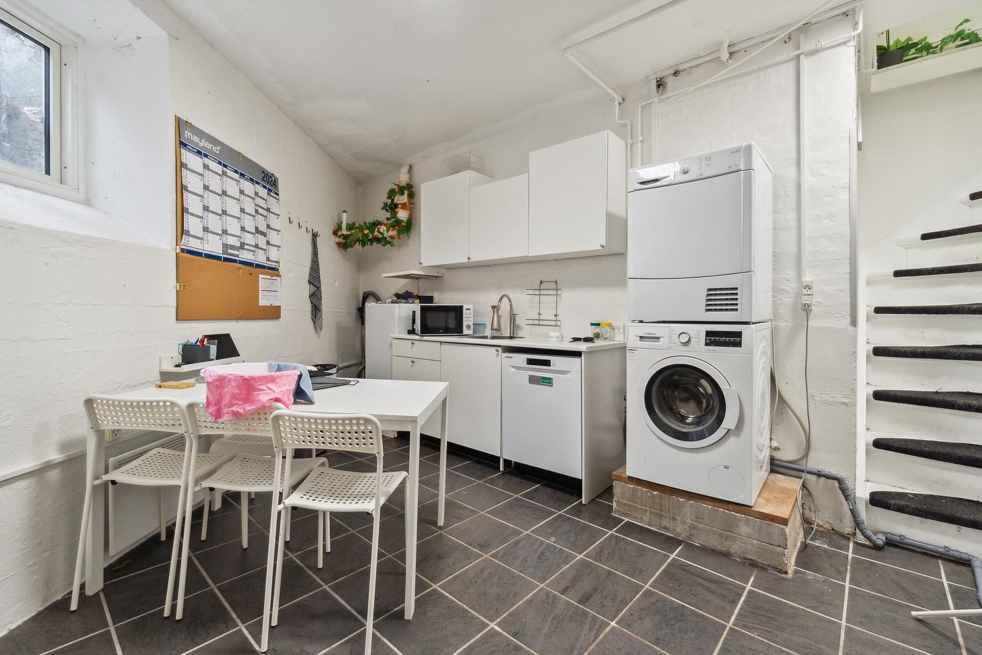 Foto af et vaskeværelse med et skrivebord og stole, samt en tørretumbler og en vaskemaskine samt en mikroovn