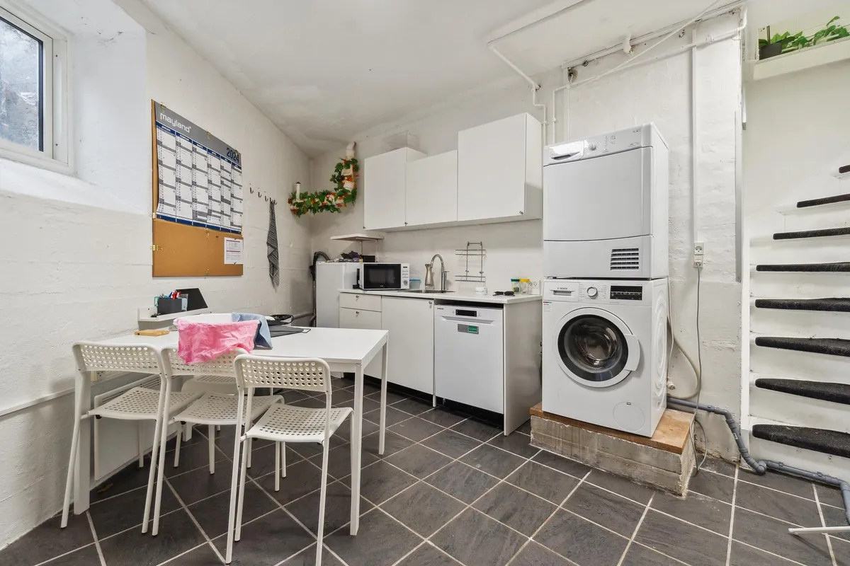 Foto af et vaskeværelse med et skrivebord og stole, samt en tørretumbler og en vaskemaskine samt en mikroovn