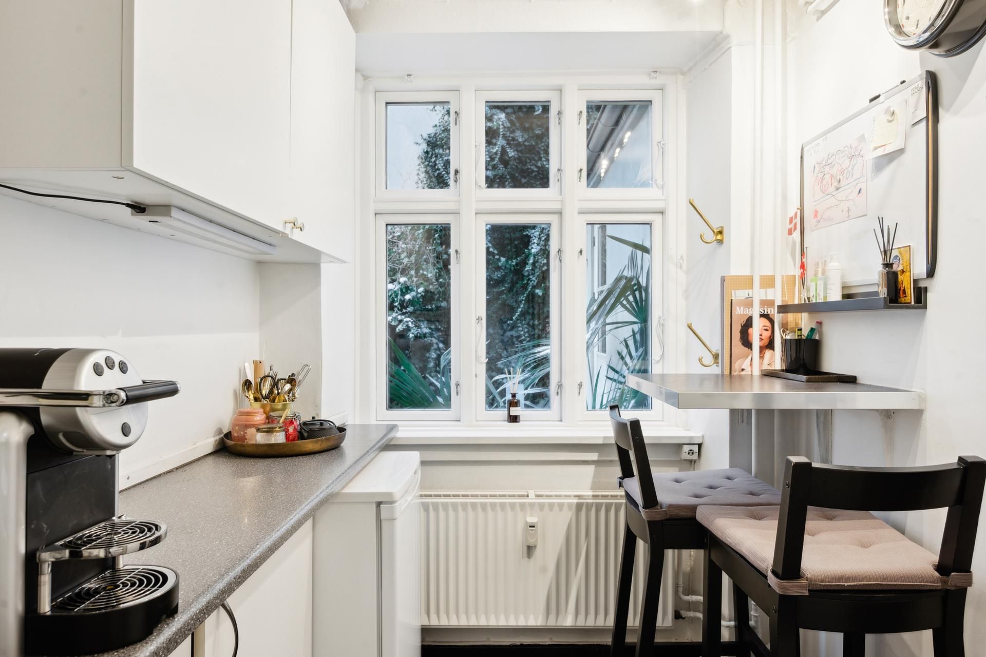 Køkken med hvide vægge, to stolpe med pudesæder og en skrivebord med aviser og lysestager på hylde og en kaffemaskine på en