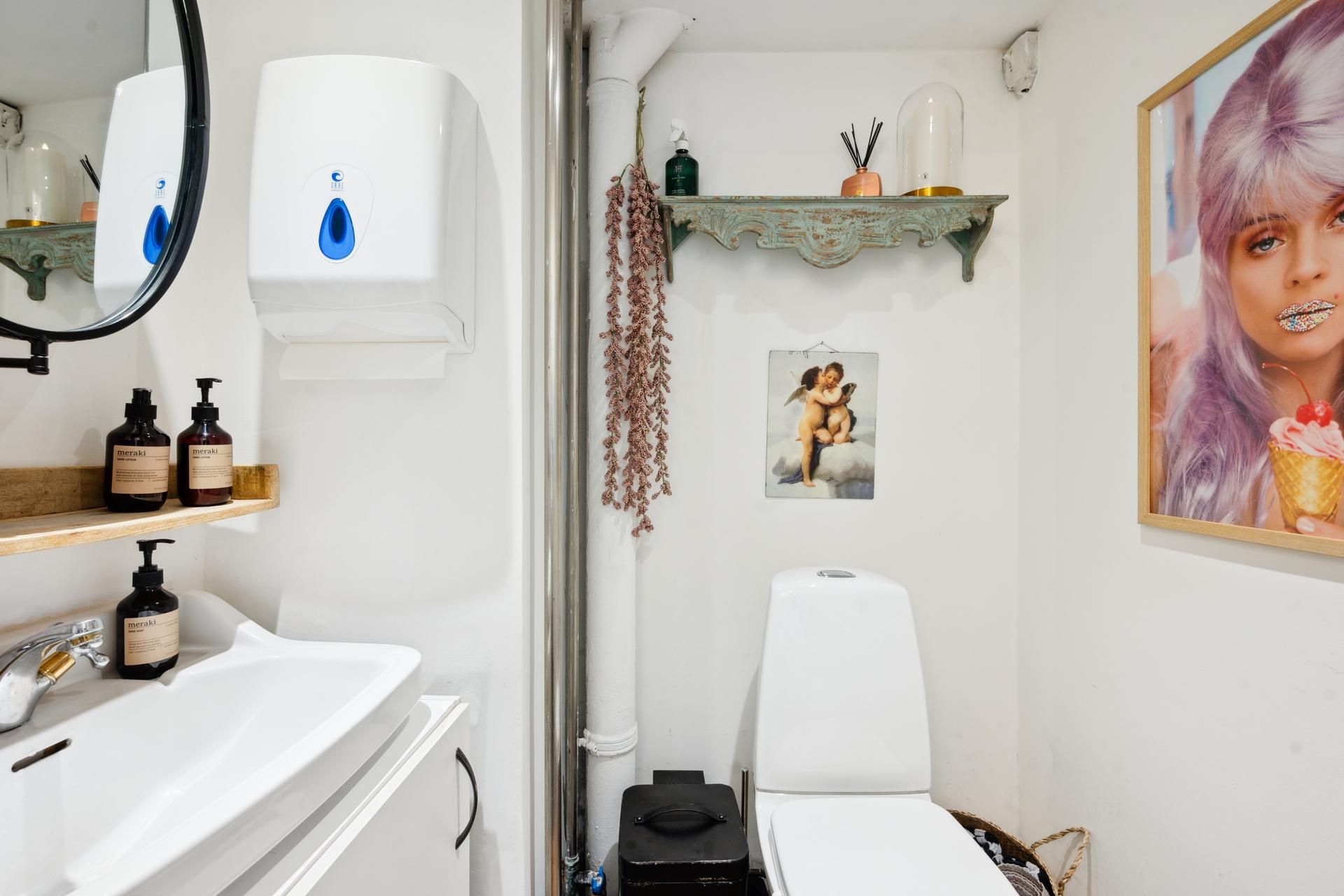 Et lille badeværelse med hvid gulv og vægge samt en håndvask med to flasker og en spejl samt et toilet og et maleri på væggen