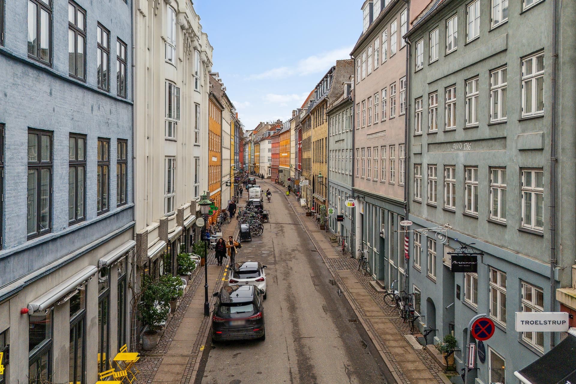 Sankt Peders Stræde i København med række af bygninger og gågade med parkerede biler og cykler, folk og butikker.